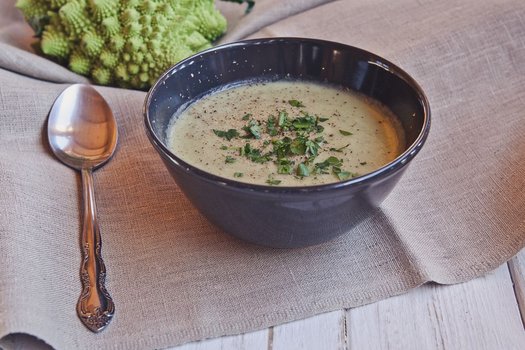 slika recepta - cvetačna juha, servirana v lepi keramični skodelici, posuta z zelišči, v ozadju cvetača romanesco